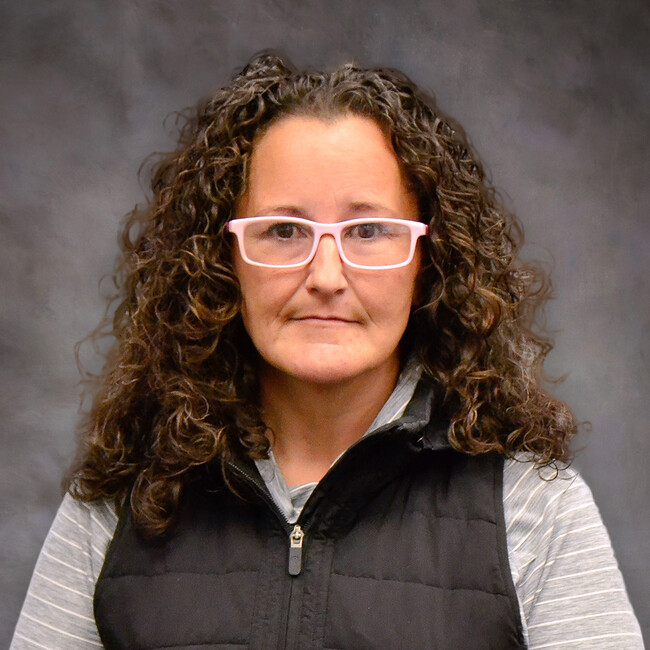 woman with curly hair wearing a black zip-up vest and glasses posing for the camera