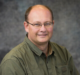 man wearing dark green button up shirt and glasses smiling for the camera